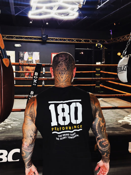 A tattooed man seen from behind wearing a black sleeveless athletic shirt with '180 PERFORMANCE' printed in white and yellow on the back, standing inside a boxing ring in a gym.